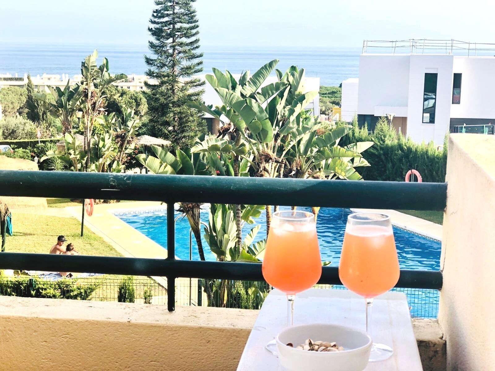 Un balcón con vista a la piscina, dos bebidas y un cuenco de frutos secos.