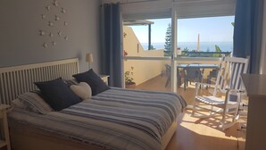 Un dormitorio con cama, almohadas, mesita de noche, lámpara, balcón con mesa y sillas, y vista al mar.