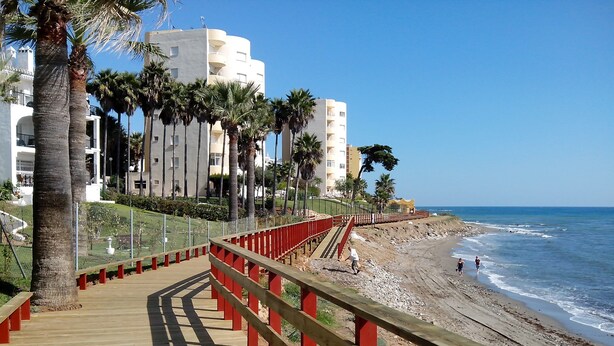 Un muelle de madera con barandillas rojas que conduce a una playa con costa rocosa y olas del océano.