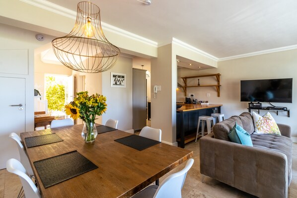 A modern living room with a wooden dining table, white chairs, a sofa with cushions, a TV mounted on the wall, and a kitchen area in the background. - Cape Town Townhouse. Panoramic Sea & Mountain Views. Beach 50m. Spectacular! (Cape Town)