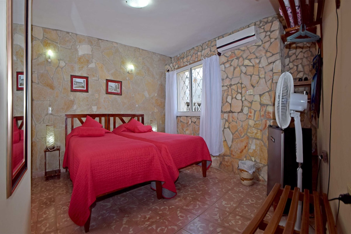 A room with two beds, stone walls, a window with curtains, a fan, and a small table with a lamp.