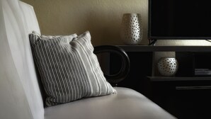 Quarto de hotel moderno com sofá branco, almofada listrada, cadeira preta e televisão sobre um móvel de madeira escura.