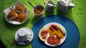 Mesa de café da manhã com pão, geleia, um bule e um prato de frutas.