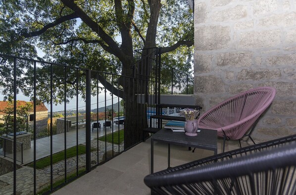 A balcony with a wicker chair, a table, and a view of a stone wall and a tree.
