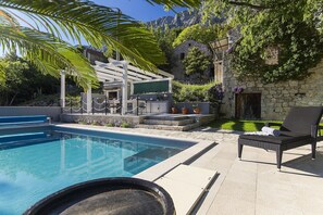 A swimming pool with a lounge area, outdoor kitchen, and stone building with greenery.