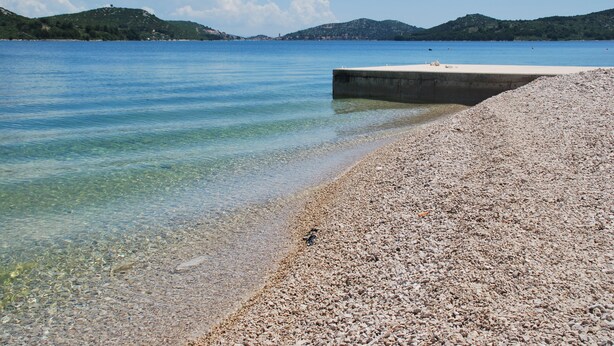 Een kiezelstrand met helder water en heuvels in de verte.