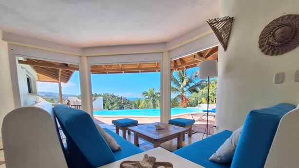 Ein Zimmer mit Blick auf einen Pool, blau gepolsterte Sitzgelegenheiten, ein Holztisch mit einer Dekoration und ein Deckenventilator.