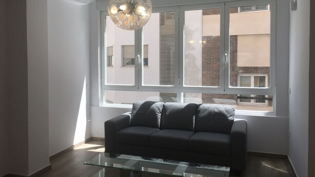 A modern living room with a glass coffee table, a dark grey sofa, and a unique circular ceiling light. Large windows provide natural light and a view of the outside. - Alicante center, beach and lively area Wifi Air-conditioning (Alacant)
