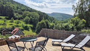 Une terrasse en bois avec une table et des chaises, donnant sur une vallée verdoyante et bordée d’une clôture en bois.