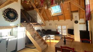 Un loft spacieux avec de hauts plafonds, des poutres en bois, une cuisine équipée d’appareils électroménagers blancs, un coin repas avec une table en bois et des chaises rouges, et un coin salon confortable avec un canapé et une cheminée.
