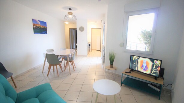 Una sala de estar con un sofá color turquesa, una mesa de centro blanca, un área de comedor con sillas, un televisor sobre un mueble bajo, y un ventanal grande.