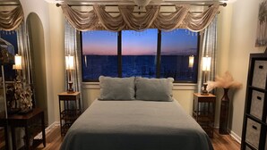 A bedroom with a large bed, two bedside tables with lamps, a view of the sea at dusk, and a decorative mirror. - Quiet Island getaway 30% off Fall/Winter stays! Private Fishing Pier, PERFECT! (Navarre)