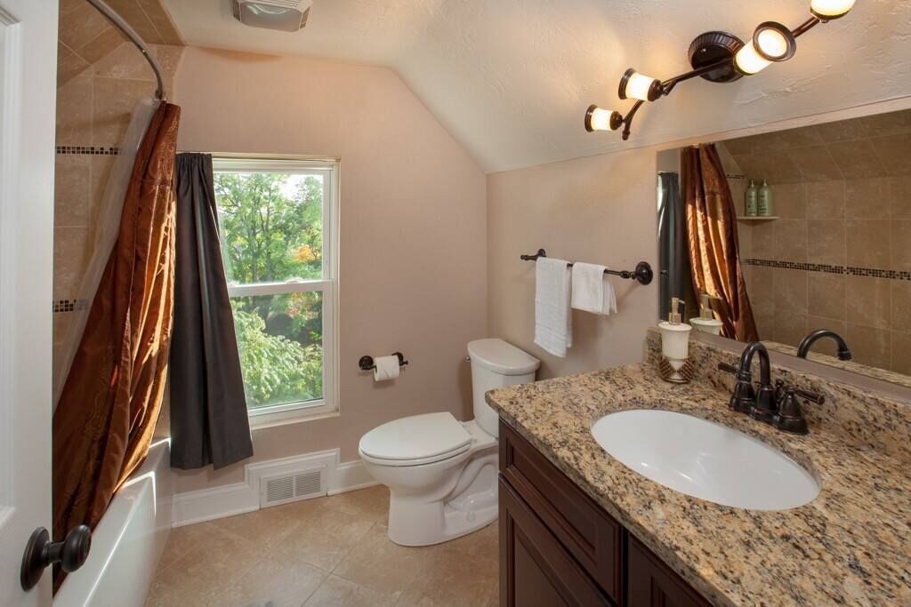 Un baño con inodoro, lavamanos y ducha. Hay una ventana con cortinas, un perchero para toallas y un espejo.