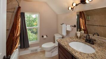 Un baño con inodoro, lavamanos y ducha. Hay una ventana con cortinas, un perchero para toallas y un espejo.