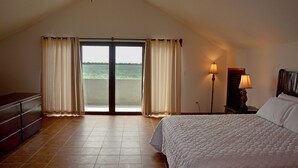 A bedroom with a bed, a dresser, a nightstand with a lamp, and a view of the sea through a sliding door.