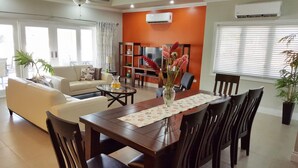 A living room with a sofa, coffee table, and dining table set with chairs. There is a TV, bookshelf, and potted plants. - Flamingo Villa in quiet & secure gated development close to airport, beaches  (Canaan)