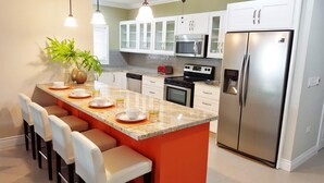 A modern kitchen with white cabinets, a stainless steel refrigerator, a microwave, and an oven. There is a bar area with stools and a plant. - Flamingo Villa in quiet & secure gated development close to airport, beaches  (Canaan)