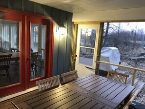 An outdoor deck with a wooden table and chairs, a red door, and a view of a snowy landscape through a window. - Get back to nature and experience lake life in style when you stay at our house (Albion)