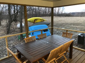A covered outdoor deck with a wooden table and chairs, a kayak, and a blue suitcase. - Get back to nature and experience lake life in style when you stay at our house (Albion)