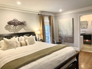 A well-lit bedroom with a large bed, a bedside table with a lamp, a wall-mounted artwork, and a view of the bathroom through an open door.