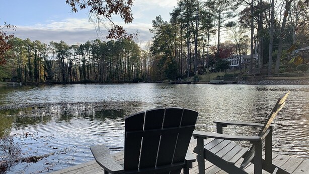 Een houten terras met twee Adirondack-stoelen gericht op een kalm meer, omgeven door hoge bomen en een gebouw in de verte.