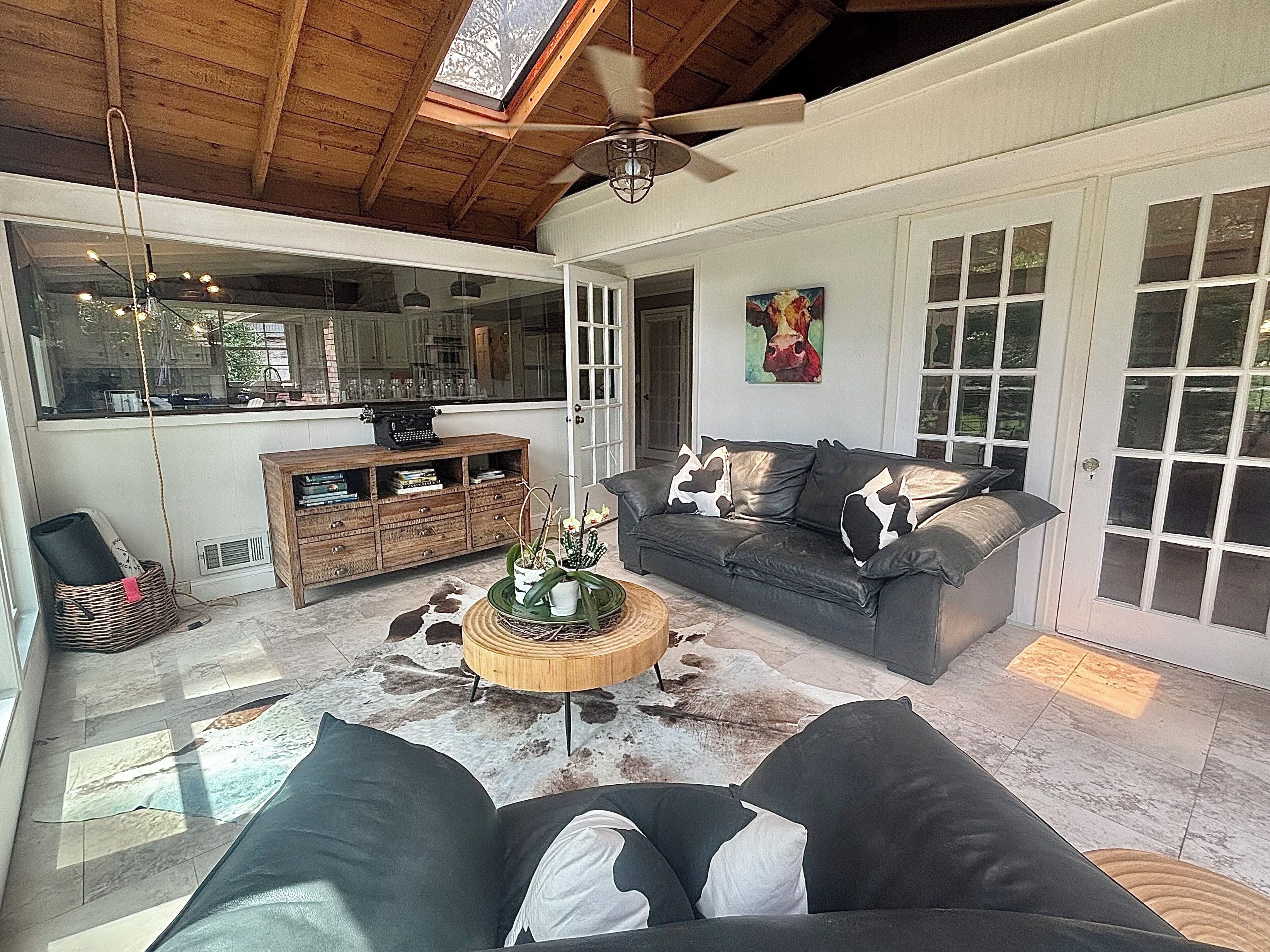 A spacious living area with a leather sofa, a wooden coffee table, and a cowhide rug. A ceiling fan and a window with a view of the outdoors are visible.