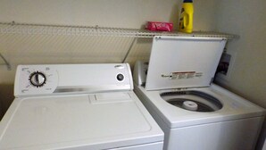 A front-loading washing machine with a control panel, a dryer, and a shelf above the dryer holding a detergent bottle and a box.