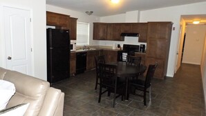 A compact kitchen with dark wooden cabinets, a black refrigerator, a dining table with four chairs, and a beige couch in the living area.