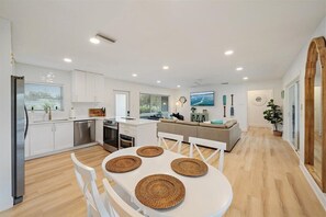 A modern kitchen with white cabinets, a dining area with a white table and chairs, and a living area with a sofa and a TV. - The Blue Conch – Family Friendly - Read About the 4th Bedroom - 3D Tour (Sarasota)