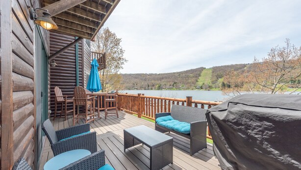 Eine Holzterrasse mit Korbmöbeln, einem überdachten Grill und Blick auf einen See und Hügel.