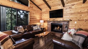 Un interior de cabaña acogedor con chimenea, sofás de cuero y un amplio ventanal con vistas al bosque.