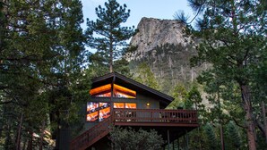 Una cabaña con terraza de madera y cerramiento de vidrio, rodeada de árboles y con la silueta de una montaña al fondo.