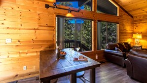 Interior de una cabaña de madera con una mesa de comedor, un sofá de cuero y grandes ventanales que ofrecen vistas al exterior.