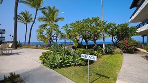 Un hôtel situé en bord de mer, avec un panneau indiquant le chemin vers la plage, des palmiers et une vue dégagée sur l’océan.