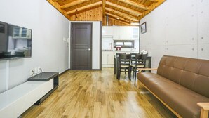 A compact living space with a wooden ceiling, a flat-screen TV mounted on the wall, a brown leather sofa, a dining table with chairs, and a kitchen area in the background. - Kyung-po Stone Valley (Gangneung)