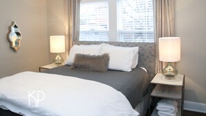 Una cama bien hecha con ropa de cama blanca y gris, dos lámparas de noche y una ventana con cortinas.