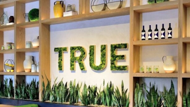 Interior de restaurante con asientos tipo cabina verdes, estantes de madera con varios objetos, y una pared con la palabra 'TRUE' en letras grandes.