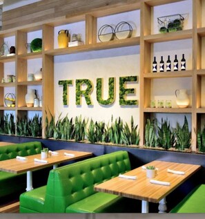 Ein Restaurantinneres mit grünen Sitzbänken, Holzregalen mit diversen Gegenständen und einer Wand, auf der das Wort 'TRUE' in großen Buchstaben steht.