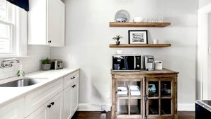 Una cocina con gabinetes blancos, un mueble de madera con puertas de vidrio, un microondas y una cafetera sobre un estante de madera.