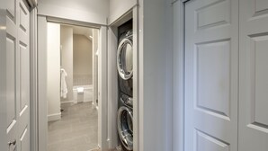 A laundry room with stacked washers and dryers, a visible door, and a hallway leading to a bathroom with a bathtub. - La Conner, Tillinghast Residence A #400 (La Conner)