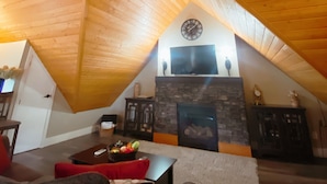A cozy attic living room with a sloped wooden ceiling, a stone fireplace, and a flat-screen TV mounted above it. - Chalet Style Suite on Peachland Acreage (Peachland)