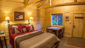Une pièce chaleureuse, aménagée dans une cabane en bois, avec un lit, deux lampes de chevet, une table et une fenêtre donnant sur un paysage verdoyant.