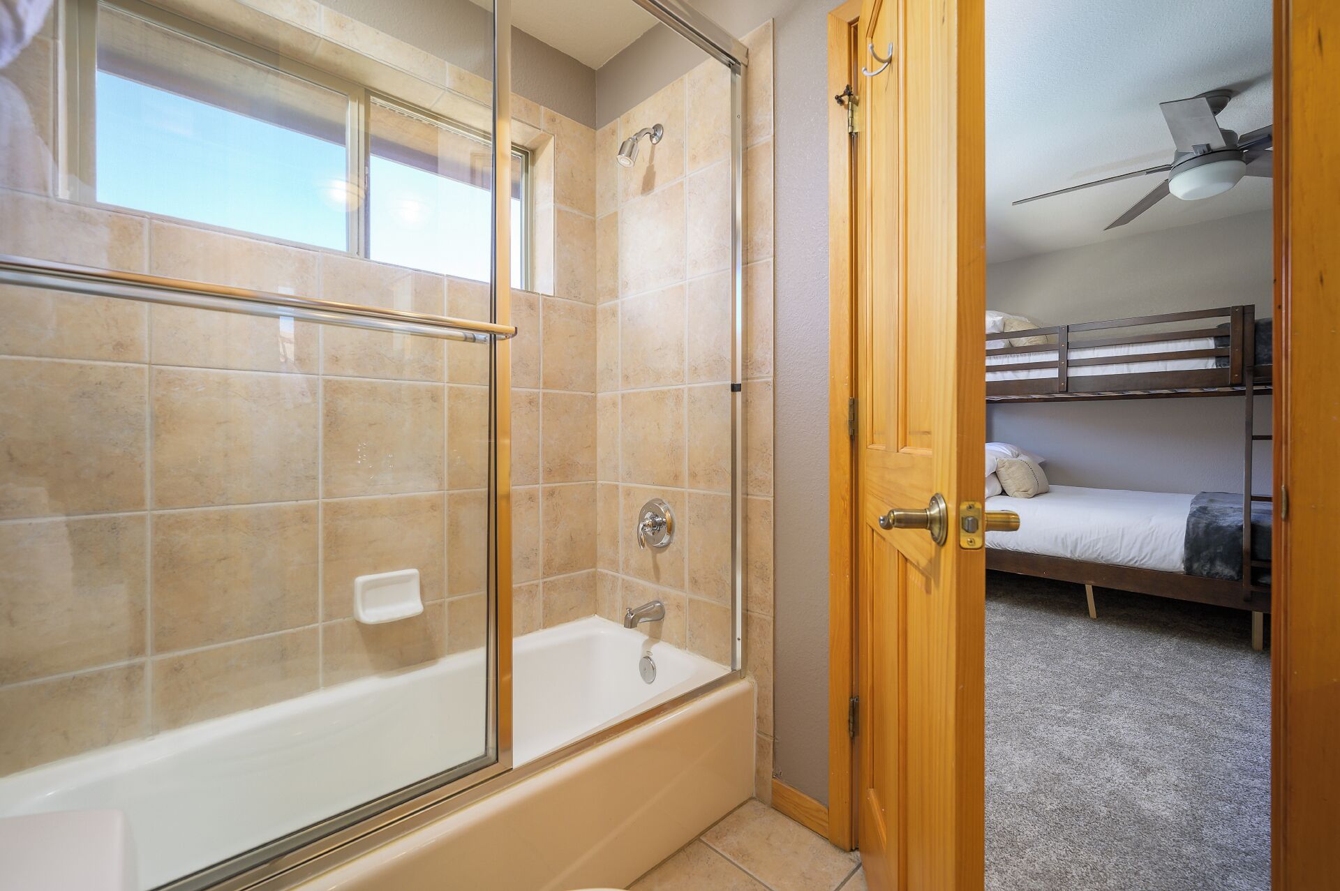 A bathroom with a glass-enclosed shower, a bathtub, and a wooden door leading to a bunk bed room.