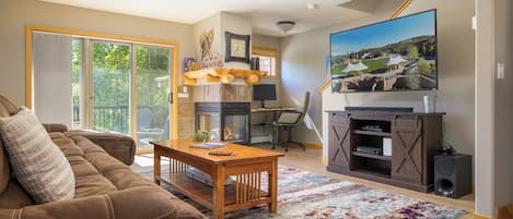 A living room with a sofa, a coffee table, a fireplace, a television, and a large window overlooking a garden.