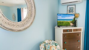Una habitación con un espejo redondo, un televisor sobre un mueble, una silla con estampado floral y una mesa de vidrio con conchas marinas.