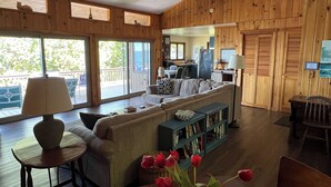 A spacious living area with a sofa, a coffee table, a bookshelf, and a dining table. There is a ceiling fan, wooden walls, and large windows.