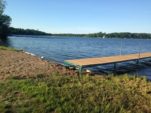 A wooden dock extending into a calm lake with a sandy shore and grassy area in the foreground. - Private Yet Middle-of-the-Action Gull Chain Home. (Nisswa)