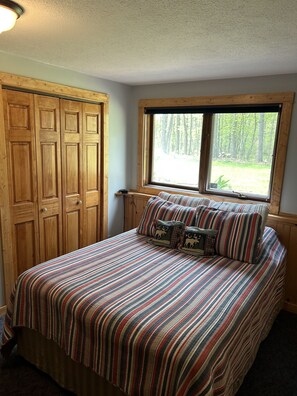 A bedroom with a large bed, striped bedding, a window with a forest view, and wooden wardrobe. - Private Yet Middle-of-the-Action Gull Chain Home. (Nisswa)