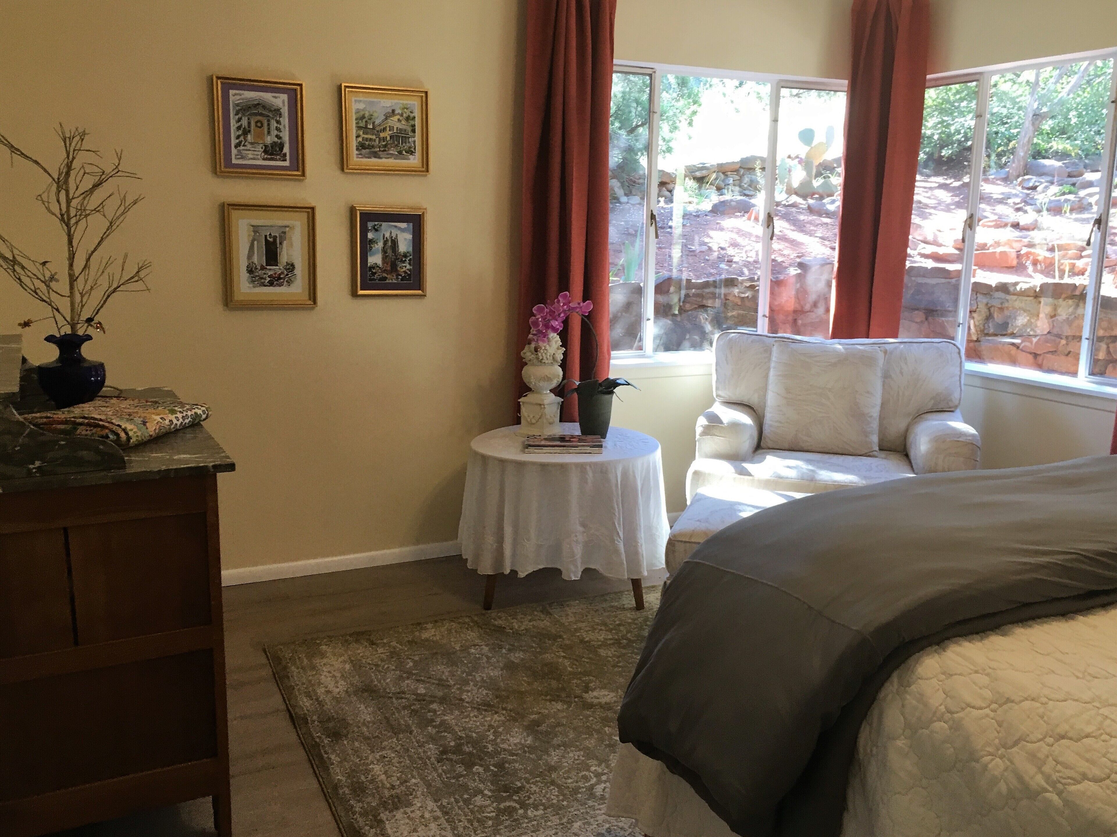 Un dormitorio con una cama, una silla, una mesita con un florero y una ventana con cortinas.