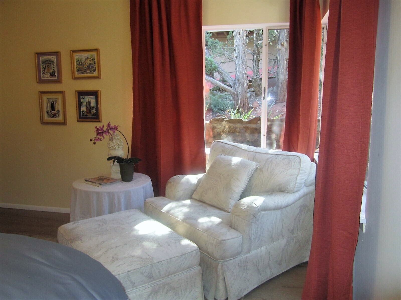 Ein Zimmer mit einer weißen Ledersofa, einem kleinen runden Tisch mit einem Topfpflanze und einem Fenster mit roten Vorhängen.